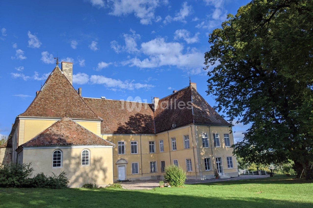 Façade d'un bâtiment historique jaune avec toit en tuiles rouges, entouré de verdure et sous un ciel bleu.