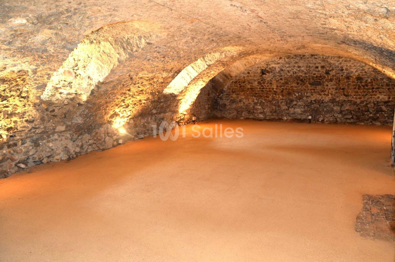 Salle voûtée en pierre avec éclairage tamisé, sol en terre battue et murs en pierre apparente.