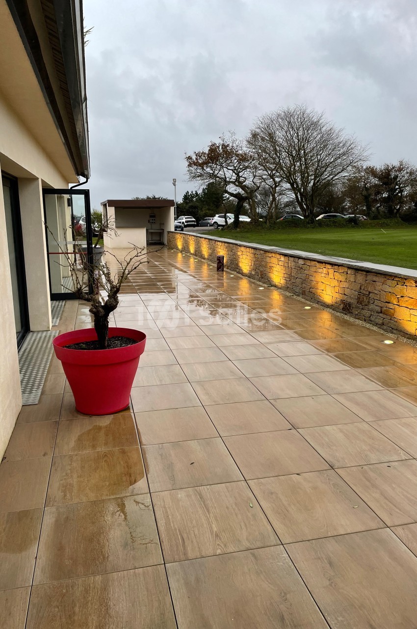 Terrasse en dalles beige mouillée par la pluie, bordée d'un mur éclairé et d'un pot rouge avec un arbuste.
