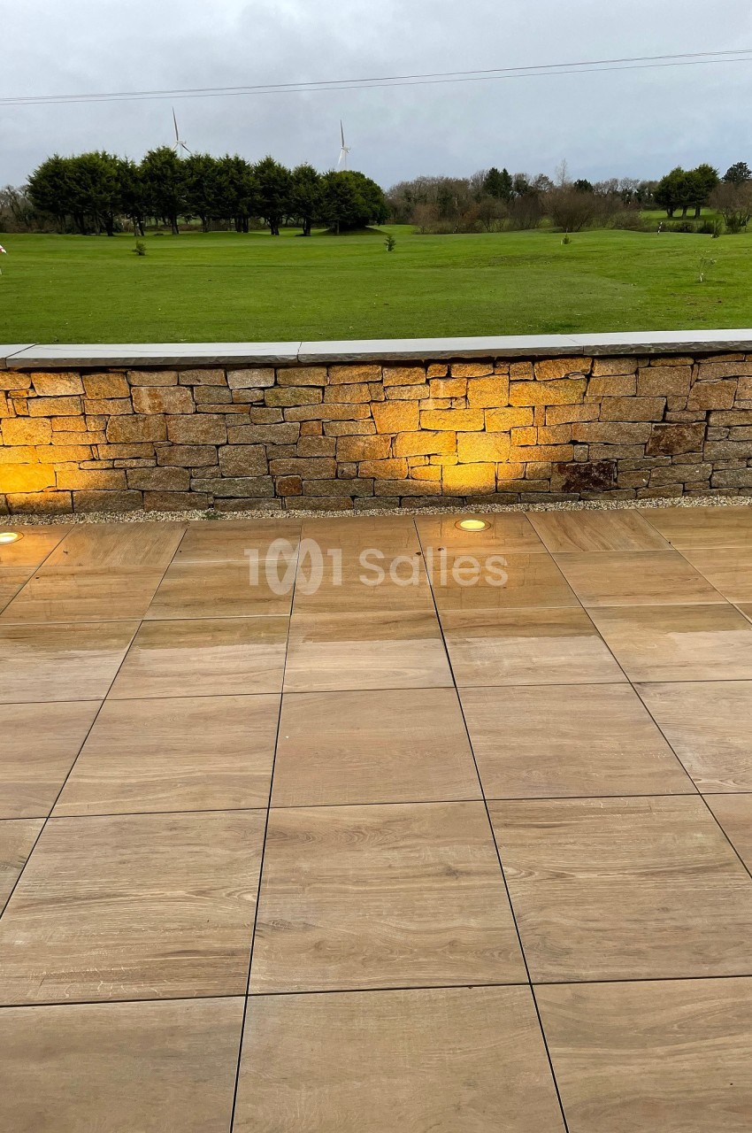 Terrasse en dalles beige clair avec éclairage au sol, mur en pierre et pelouse arborée en arrière-plan.