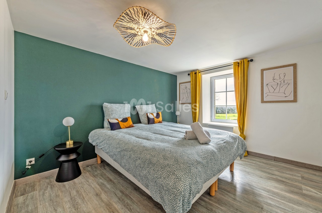Chambre lumineuse avec lit double, mur vert, rideaux jaunes, parquet clair et lampe design au plafond.