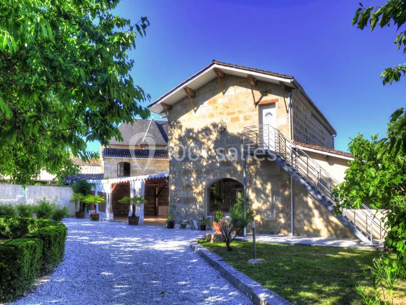 Maison en pierre avec escalier extérieur, entourée de verdure et d'une allée gravillonnée sous un ciel bleu.
