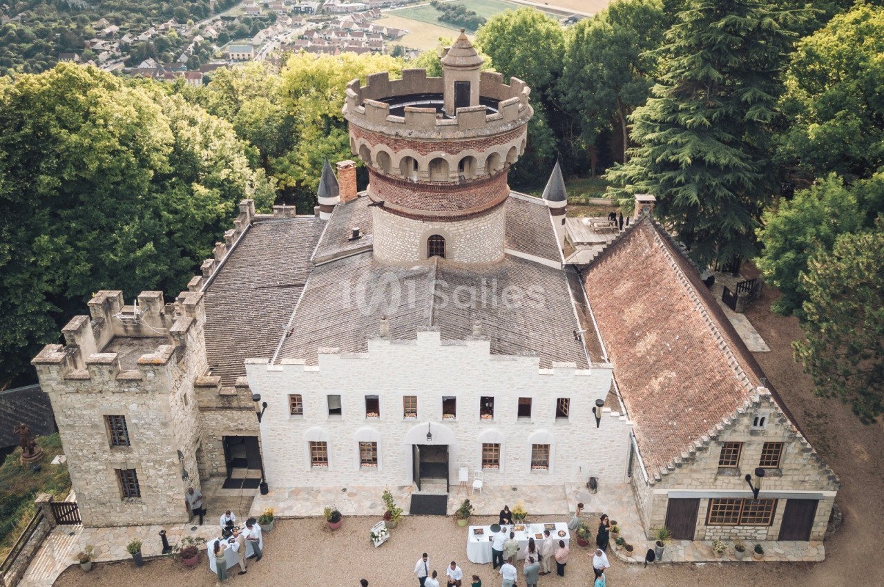 Vue aérienne d'un château entouré de verdure, avec des invités rassemblés dans la cour pour un événement.