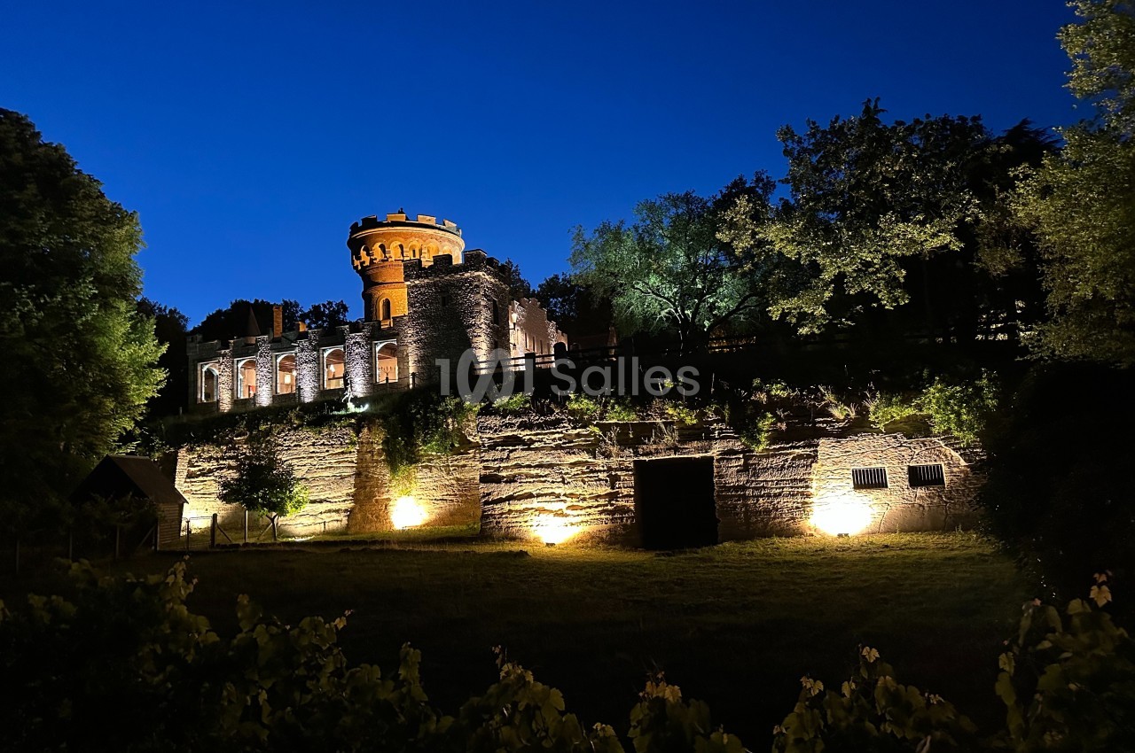 Château illuminé la nuit, entouré d'arbres et surplombant un jardin sombre.