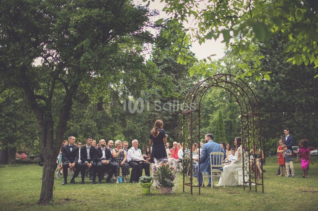 Cérémonie de mariage en plein air avec des invités assis, un couple marié et une officiante sous une arche fleurie.