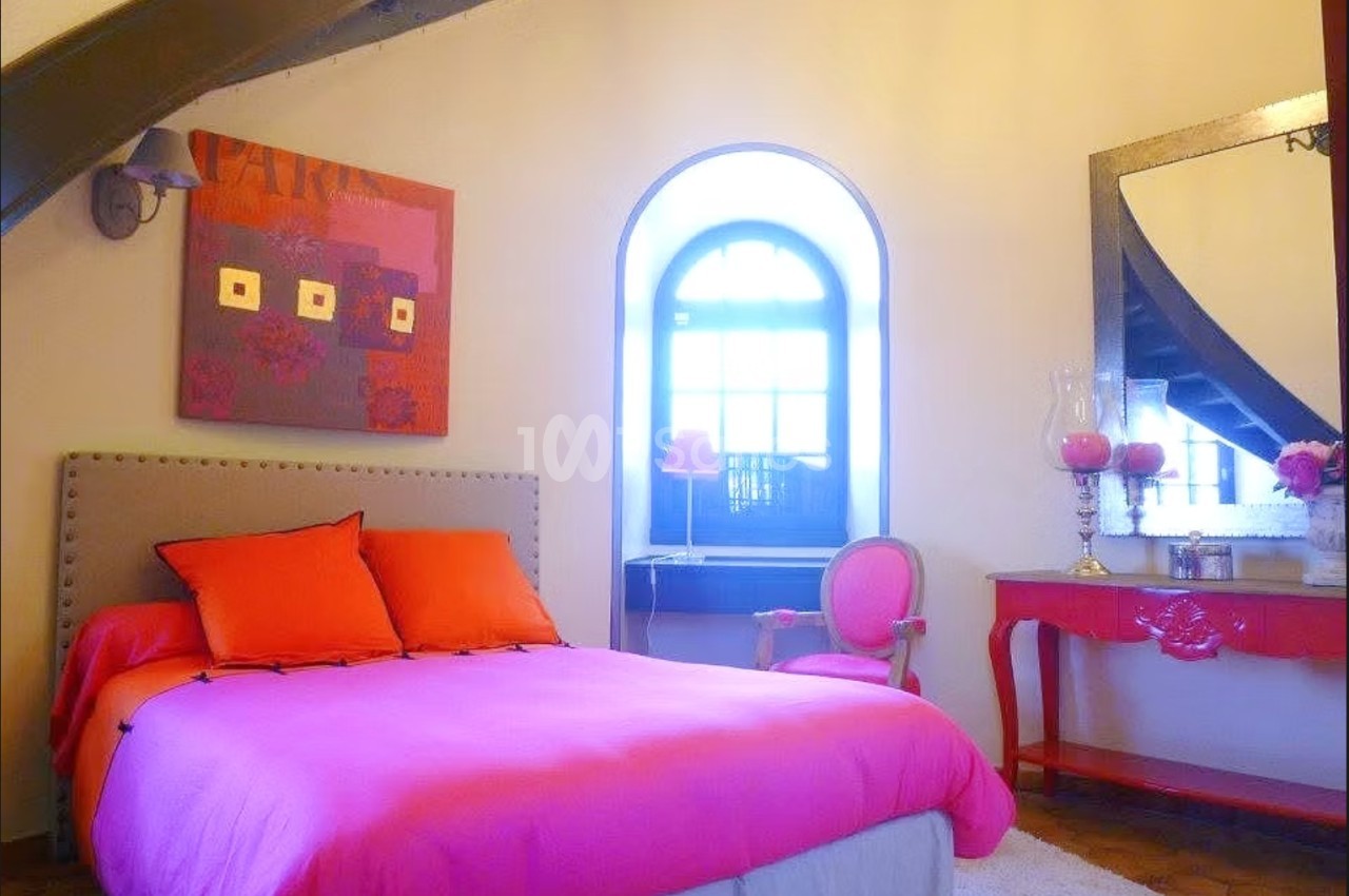 Chambre lumineuse avec lit rose, fauteuil assorti, bureau rouge et grande fenêtre voûtée laissant entrer la lumière.