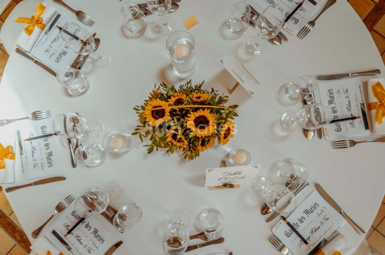Table ronde décorée avec un bouquet de tournesols au centre, entourée de couverts et de menus disposés pour des invités.