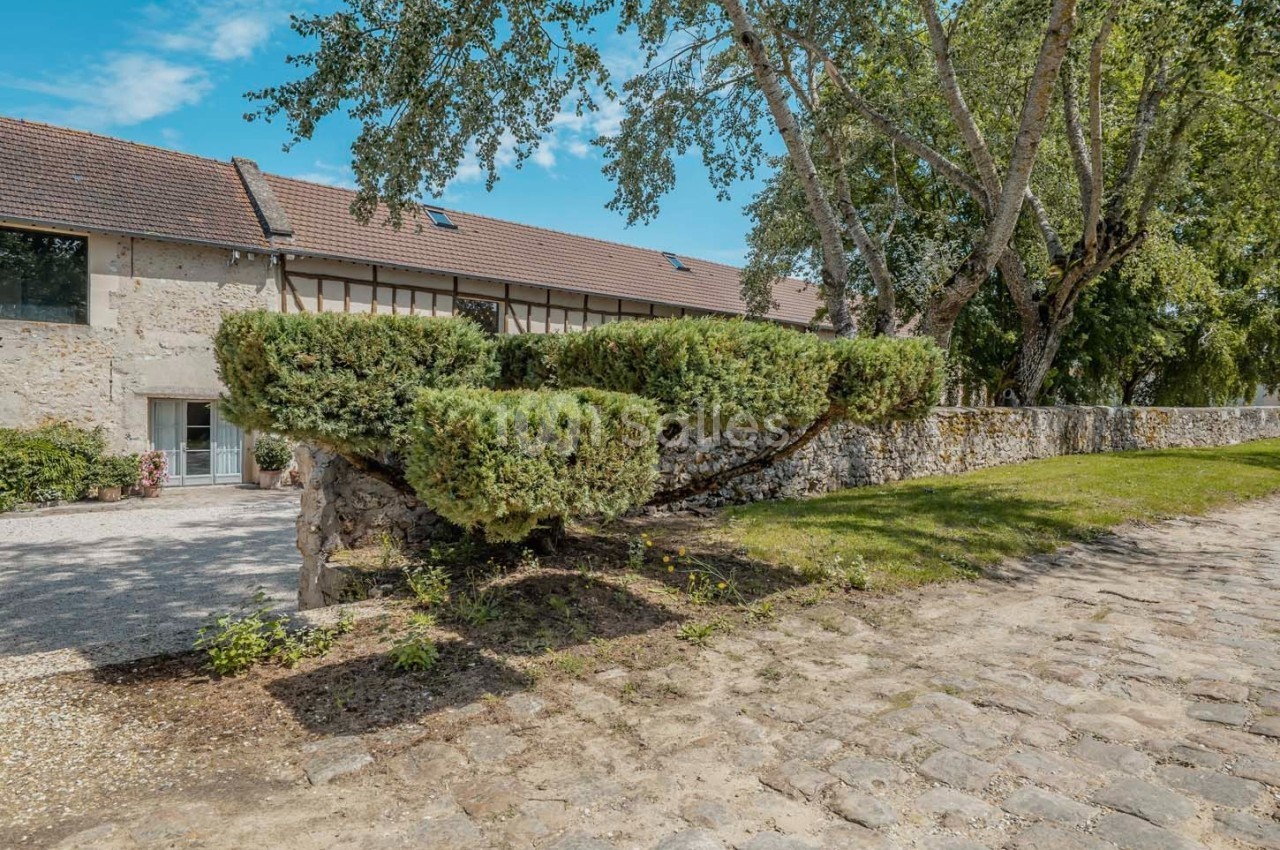Façade en pierre d'une maison avec toit en tuiles, haies taillées, allée pavée et arbres en arrière-plan.