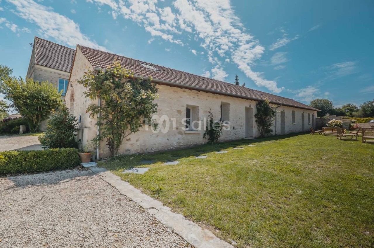 Façade d'une maison en pierre avec toit en tuiles, entourée de verdure et d'un ciel bleu parsemé de nuages.