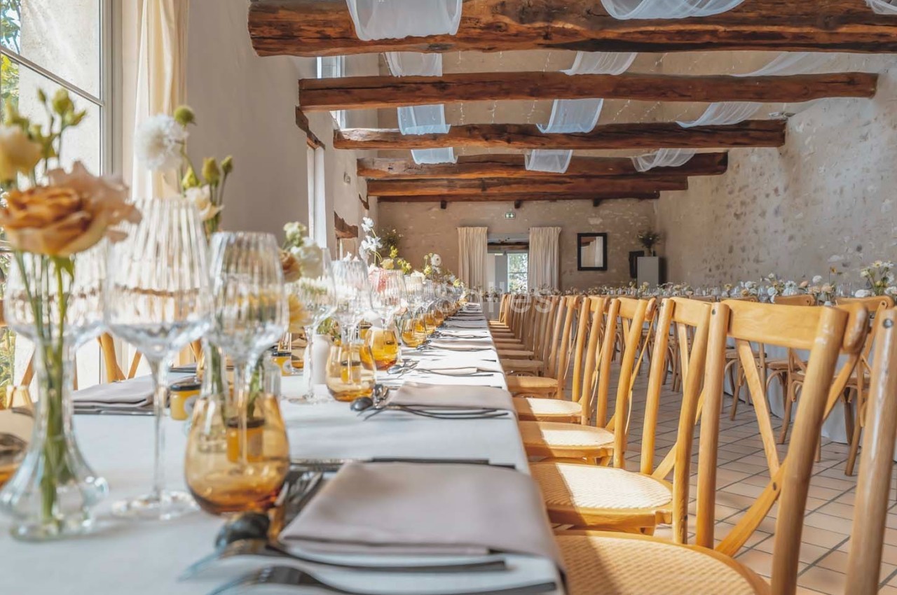 Salle de réception lumineuse avec une longue table dressée, décorée de fleurs et de verres élégants.