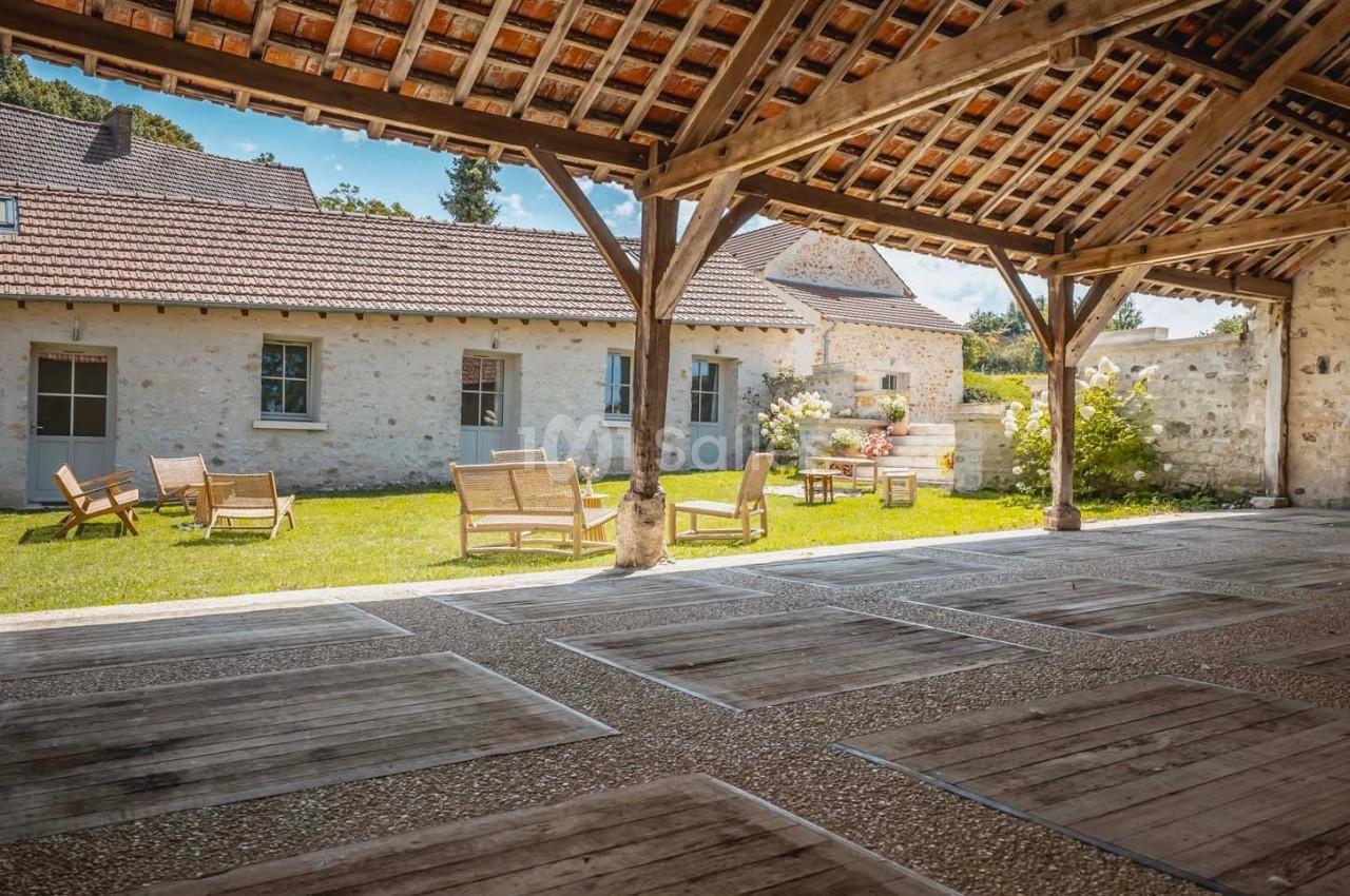 Cour intérieure avec pelouse, mobilier de jardin en bois et bâtiments en pierre sous un auvent en bois.