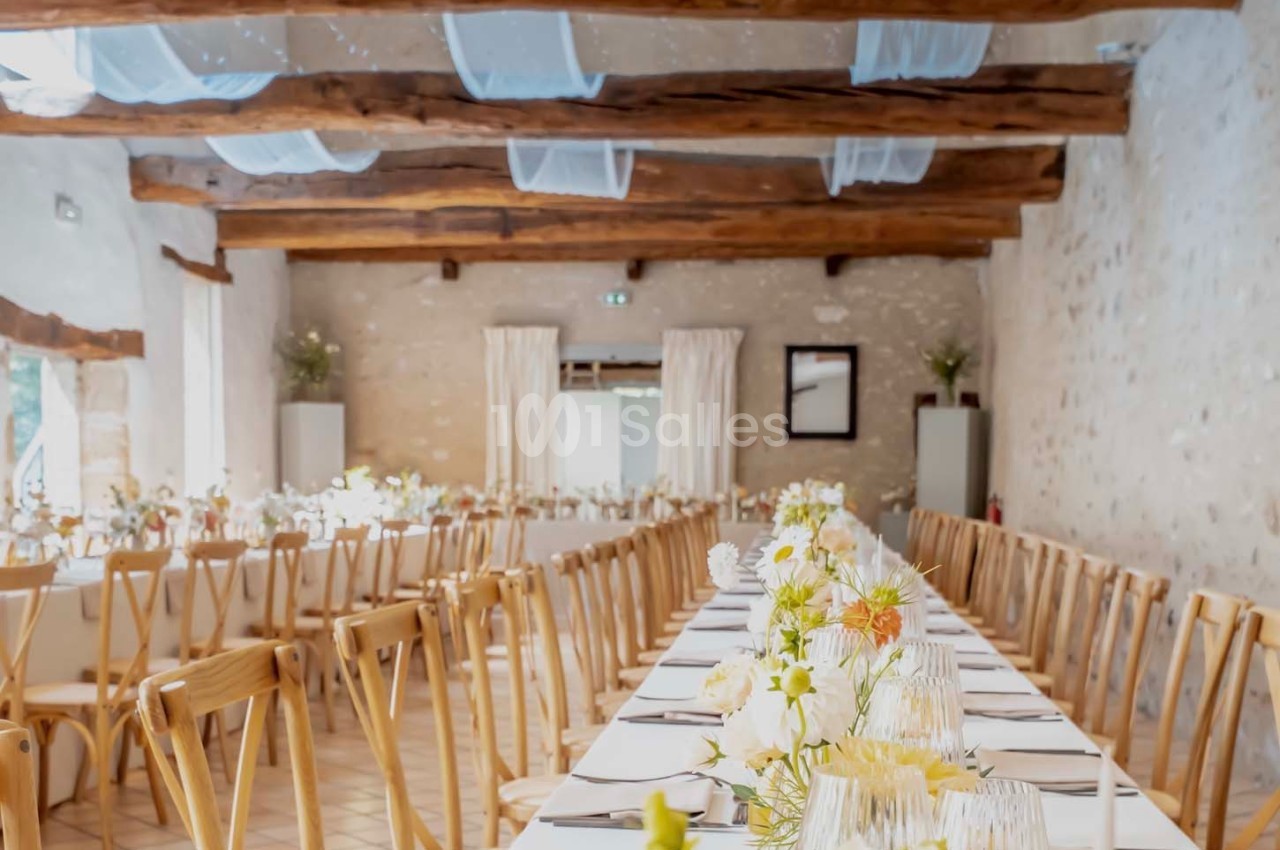 Salle de réception lumineuse avec tables alignées, nappes blanches, chaises en bois et décoration florale.