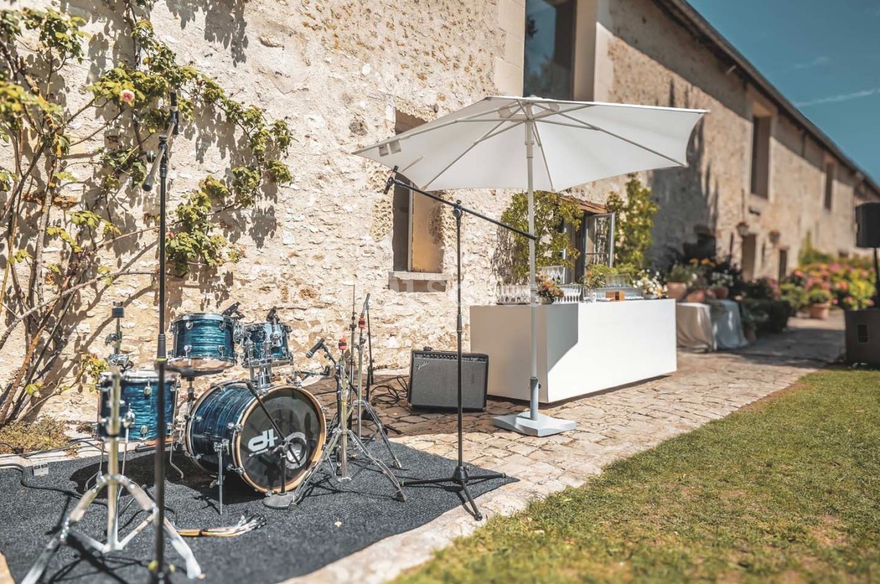 Batterie et micros installés en extérieur sur une terrasse pavée près d'un bar sous un parasol, devant un mur en pierre.