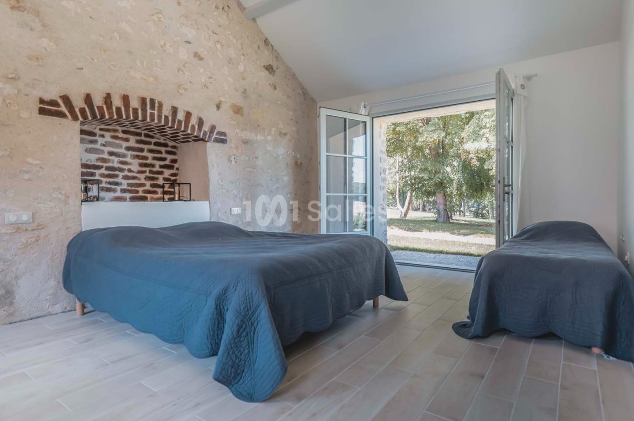 Chambre lumineuse avec murs en pierre, deux lits recouverts de couettes bleues et porte-fenêtre donnant sur un jardin.