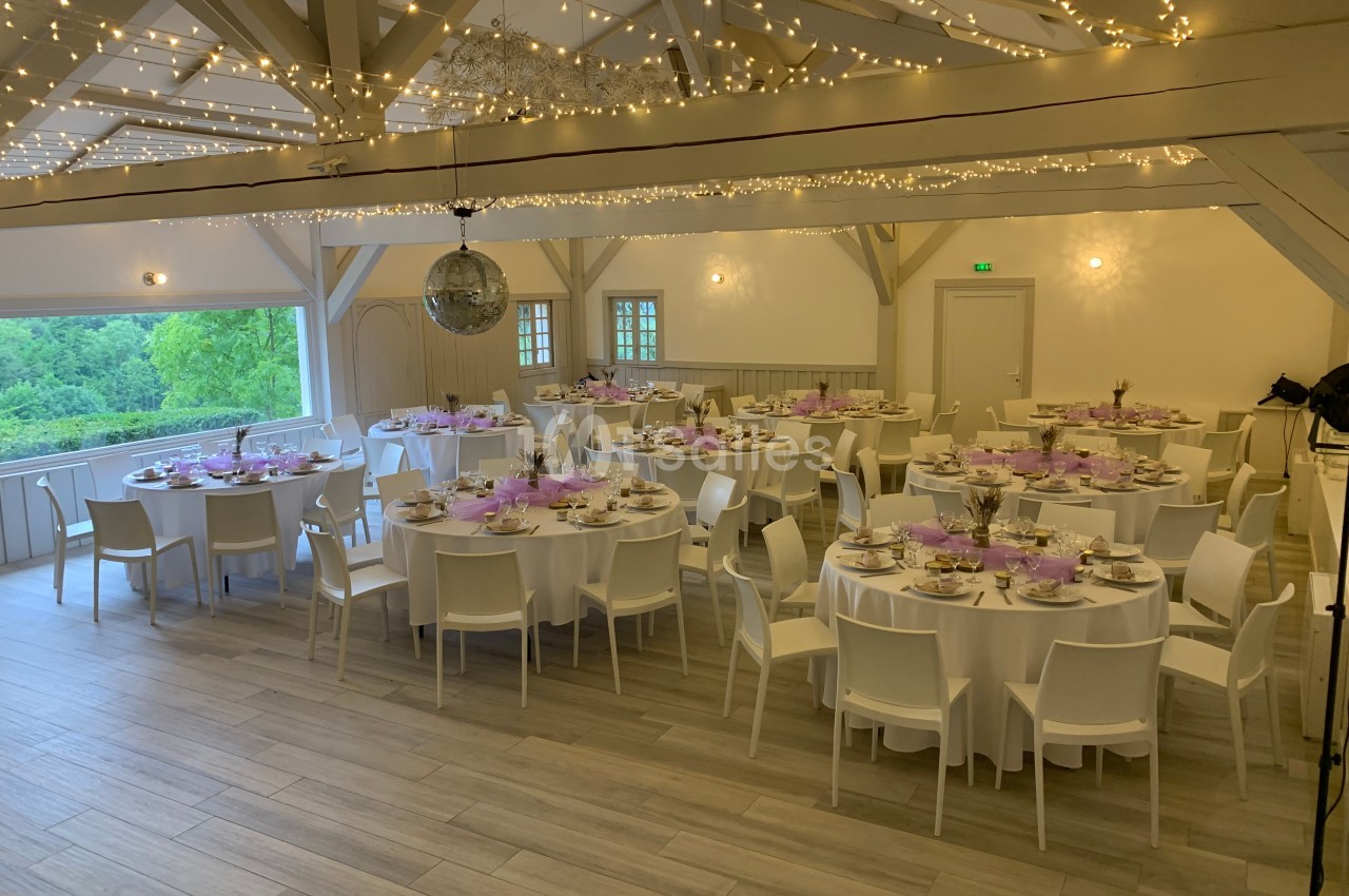 Salle de réception décorée avec des tables rondes dressées, guirlandes lumineuses suspendues et vue sur un paysage verdoyant.