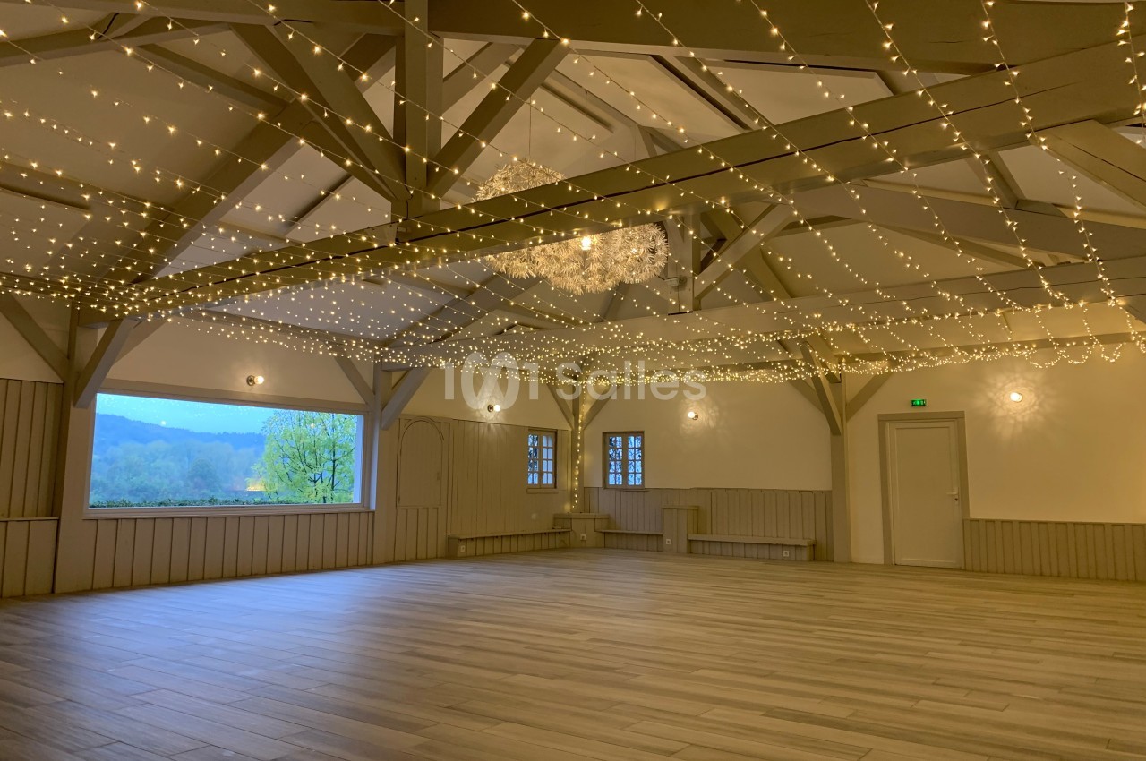Salle spacieuse avec parquet clair, poutres apparentes et guirlandes lumineuses suspendues au plafond.