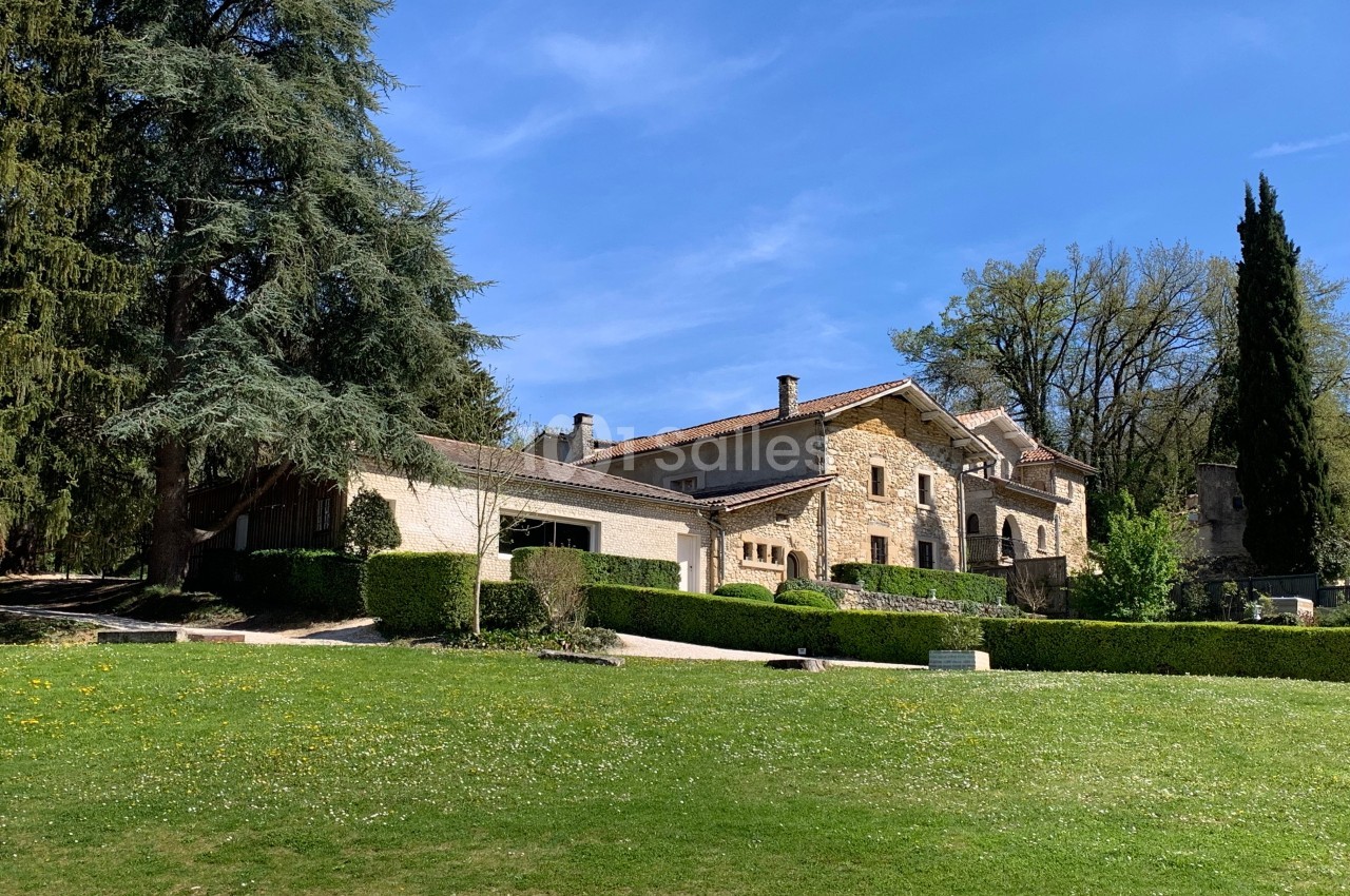 Grande maison en pierre entourée d'arbres et de pelouses, sous un ciel bleu dégagé.