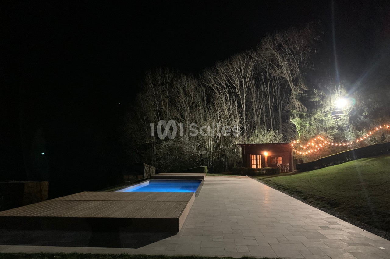 Piscine éclairée la nuit avec une terrasse en bois, un chalet illuminé et des arbres en arrière-plan.