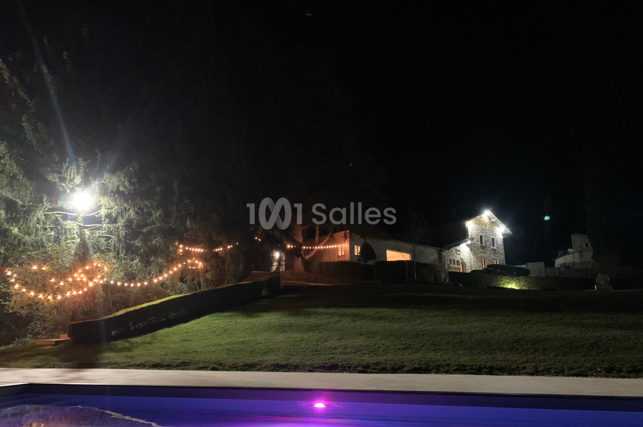 Jardin éclairé de nuit avec guirlandes lumineuses, maison en arrière-plan et piscine illuminée au premier plan.