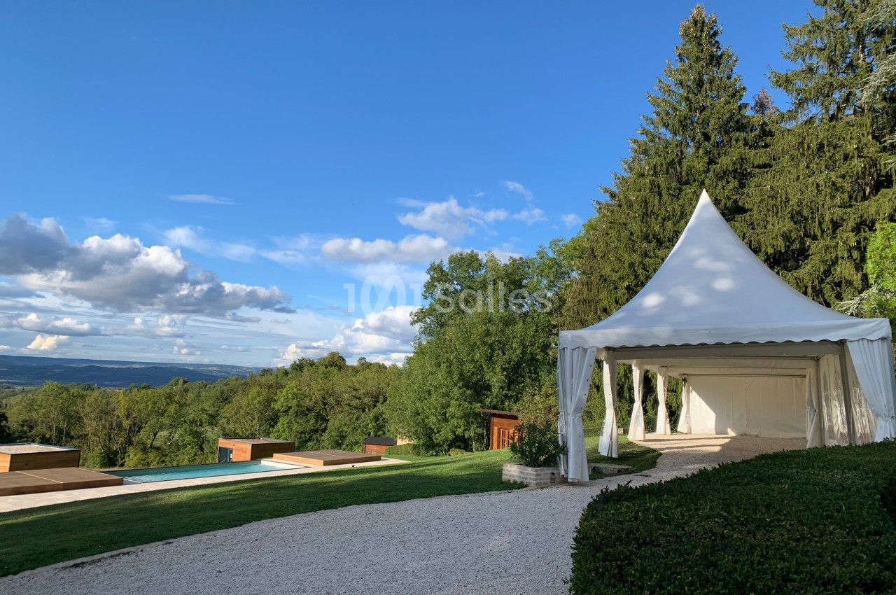 Tente blanche installée dans un jardin avec vue sur une piscine, des arbres et un paysage vallonné sous un ciel bleu.