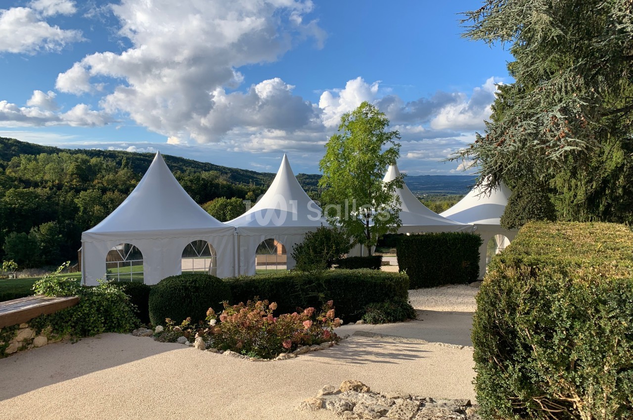 Tentes blanches pointues installées dans un jardin paysager avec vue sur une vallée boisée sous un ciel partiellement…