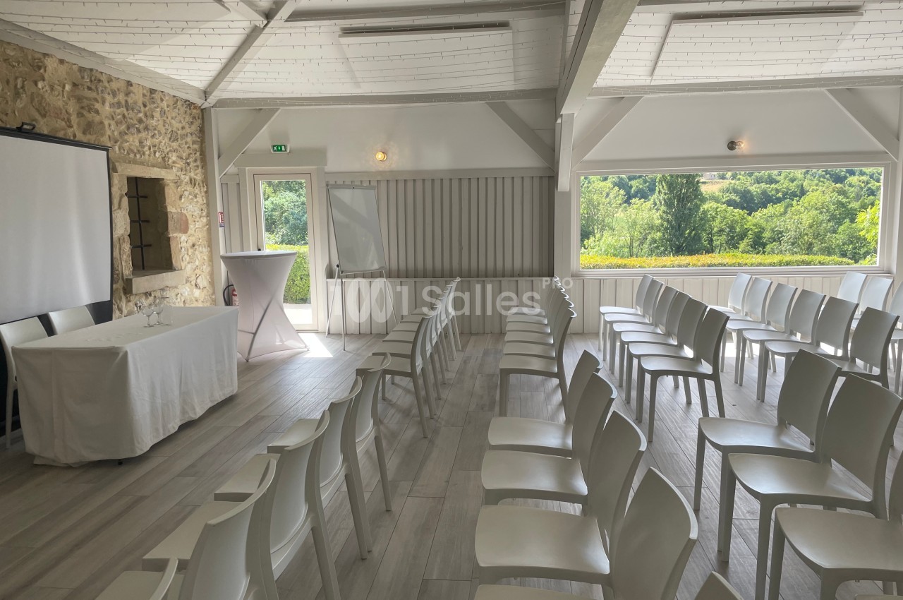 Salle lumineuse avec chaises blanches alignées, écran de projection et vue sur un paysage verdoyant à travers une grande…
