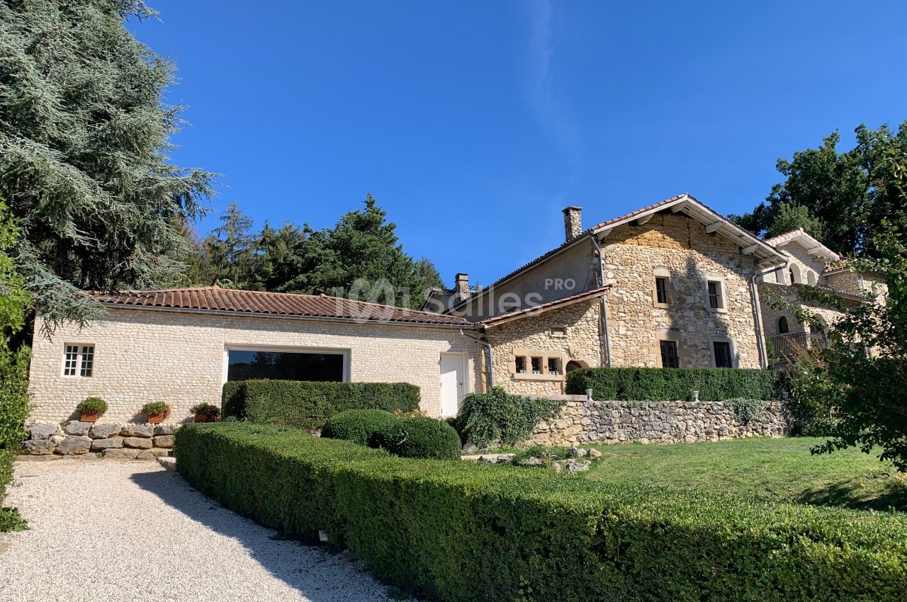 Maison en pierre avec toit en tuiles, entourée de haies taillées, d'arbres et d'une allée en gravier sous un ciel bleu.