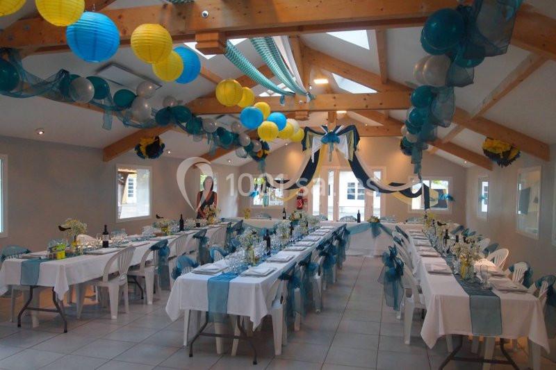 Salle décorée pour une réception avec tables dressées, nappes blanches, chemins de table bleus et ballons suspendus.