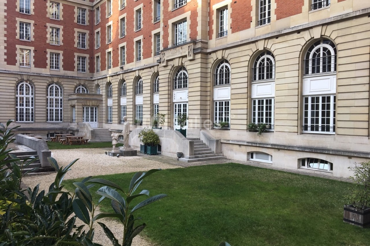 Cour intérieure d'un bâtiment en briques et pierre, avec pelouse, bancs, tables et grandes fenêtres en arc.