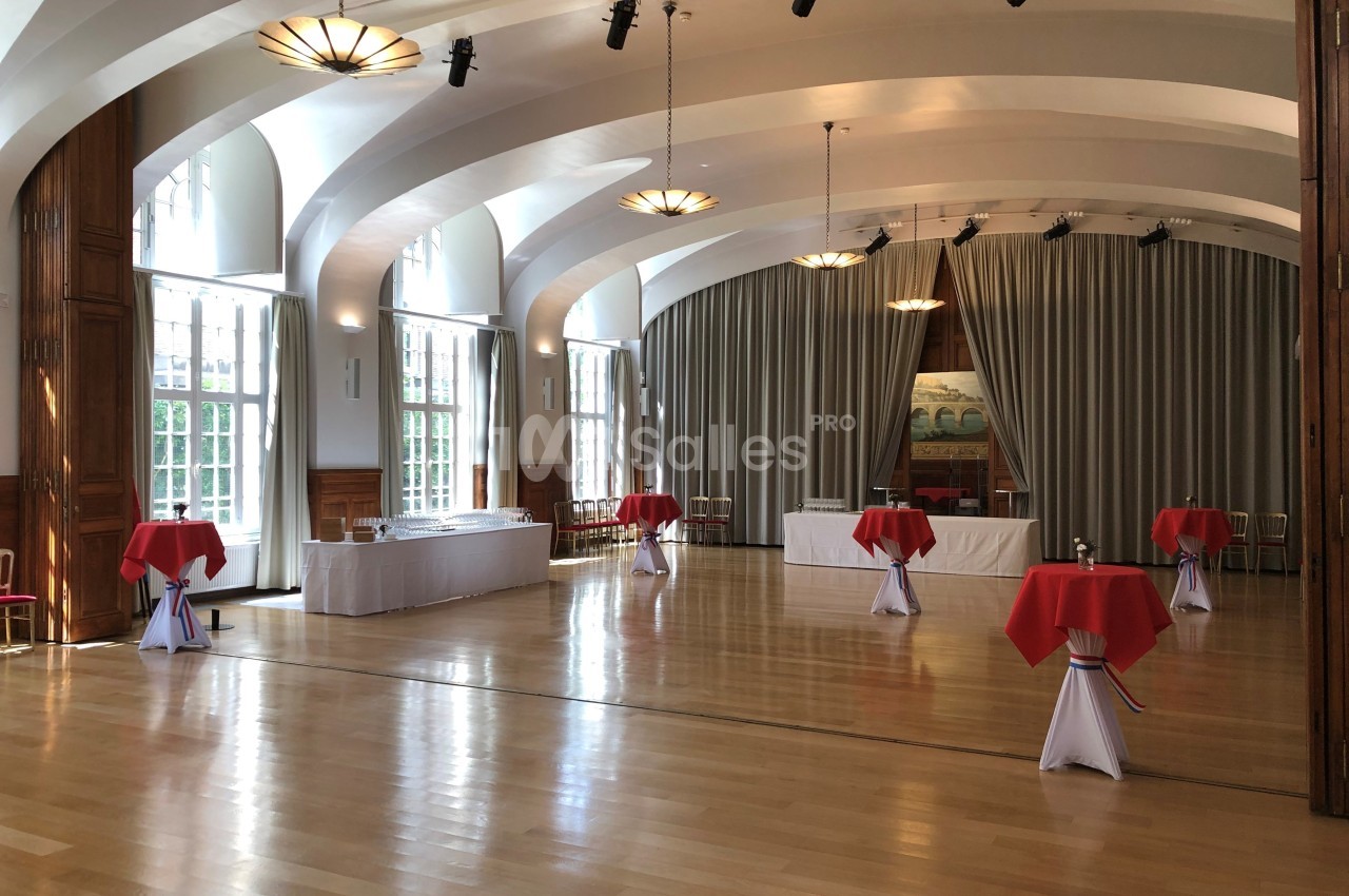 Salle de réception lumineuse avec parquet, tables hautes nappées de rouge et grandes fenêtres donnant sur l'extérieur.