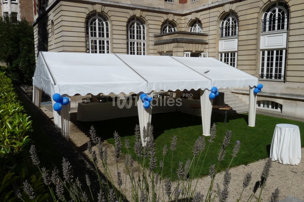 Tente blanche installée dans une cour avec des ballons bleus attachés aux poteaux et des tables en dessous.