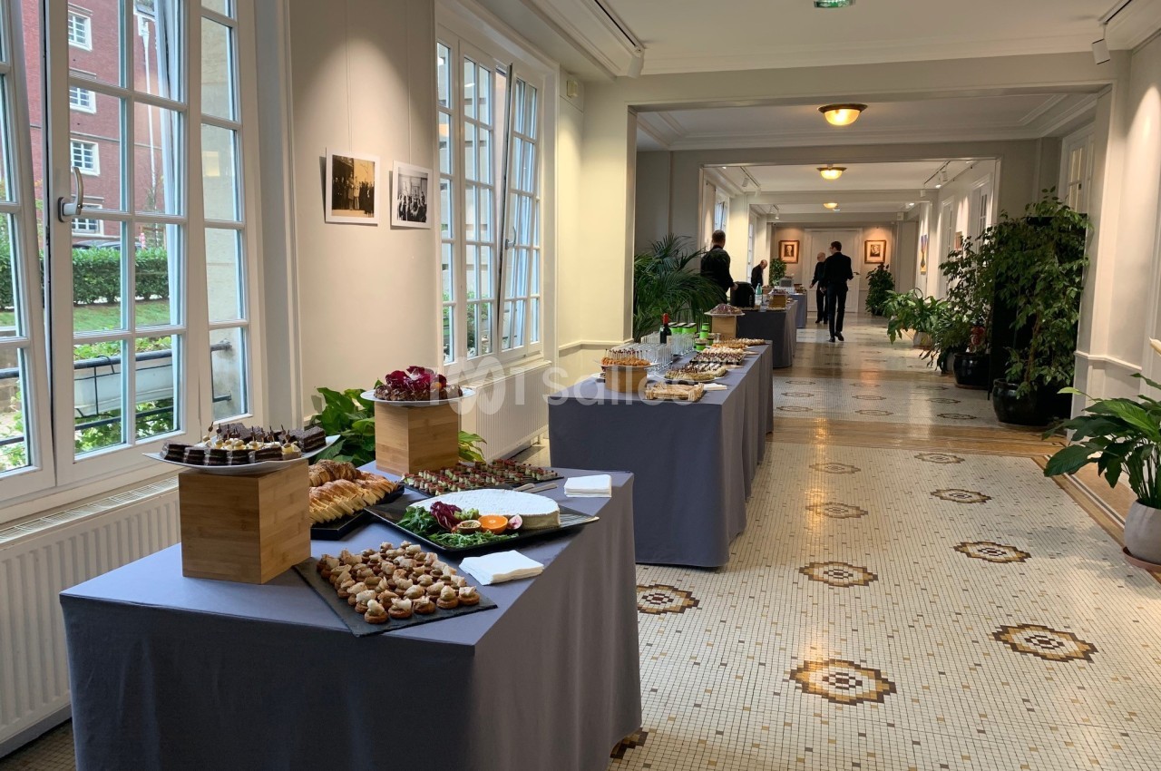 Tables dressées avec des buffets de nourriture dans un couloir lumineux, décoré de plantes et de carrelage ancien.