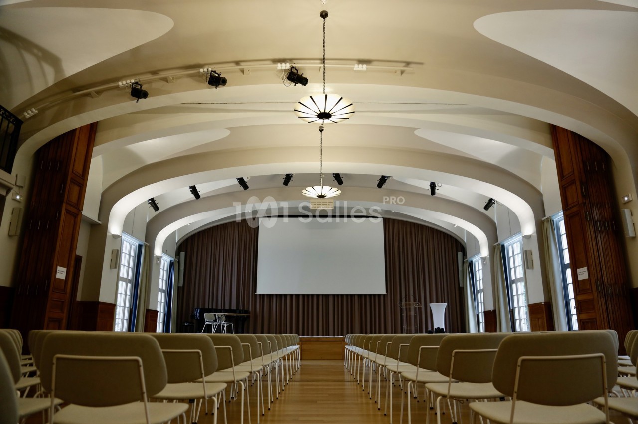 Salle de conférence lumineuse avec rangées de chaises, scène équipée d'un écran, rideaux et éclairage suspendu.