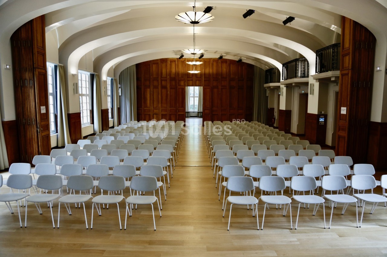 Salle de conférence lumineuse avec rangées de chaises blanches alignées sur un sol en bois clair.