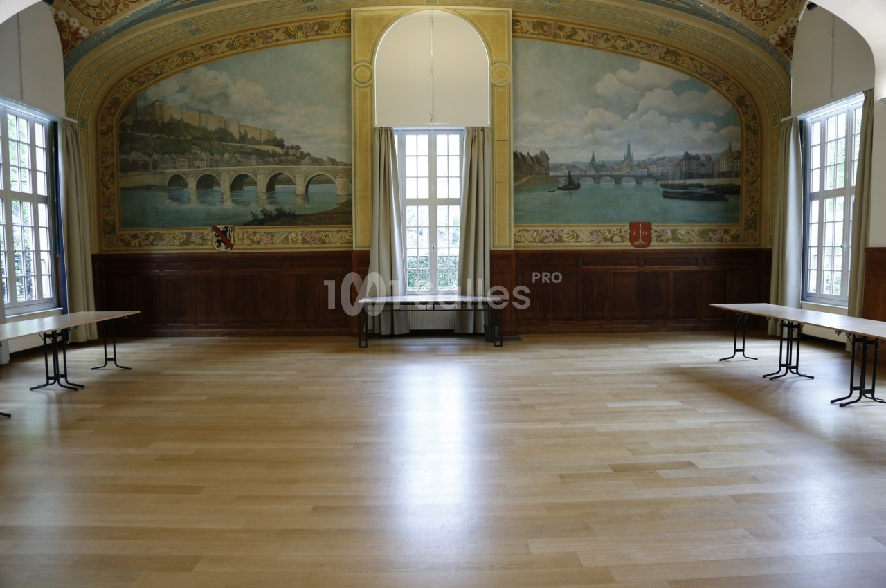 Salle vide avec parquet clair, murs ornés de fresques représentant un paysage fluvial et grandes fenêtres laissant entrer la…