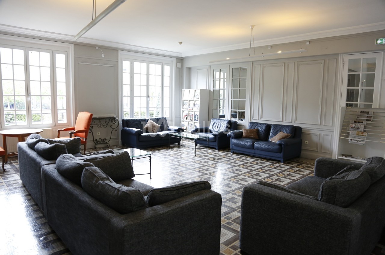 Salle lumineuse avec canapés gris et bleus, fauteuil orange, grandes fenêtres et sol en carreaux.