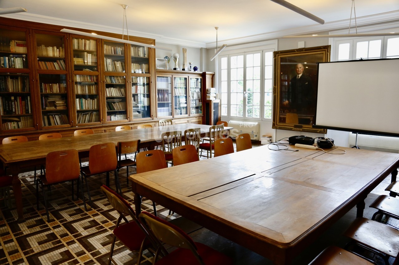 Salle de réunion avec tables en bois, chaises, bibliothèque remplie de livres et un écran de projection.