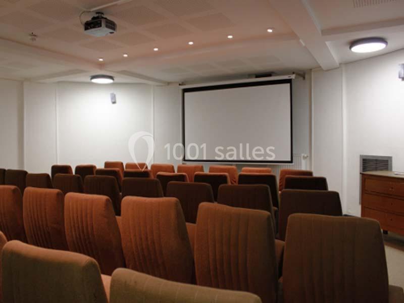 Salle de projection avec rangées de fauteuils en tissu marron, écran blanc et éclairage au plafond.