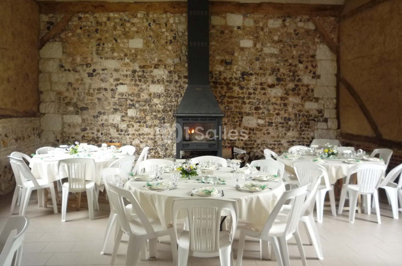 Salle rustique avec murs en pierre, cheminée allumée et tables rondes dressées pour un repas, entourées de chaises blanches.