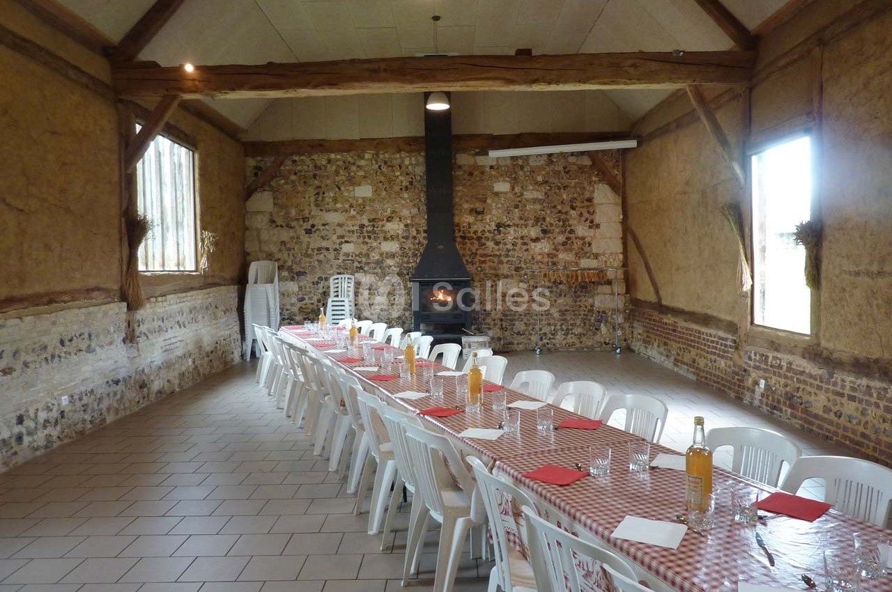 Grande salle rustique avec murs en pierre, cheminée allumée et longues tables dressées avec nappes à carreaux rouges.