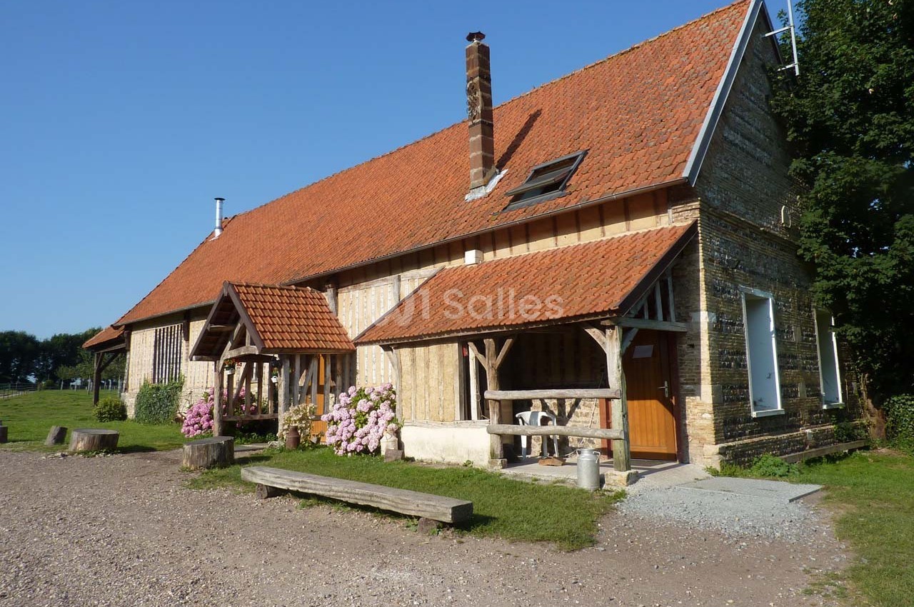 Maison en briques et bois avec toit en tuiles rouges, entourée de verdure et ornée de fleurs roses.