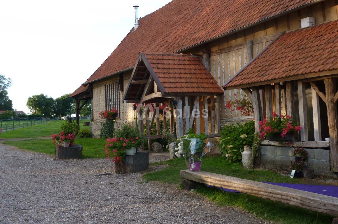 Ferme traditionnelle en bois avec toit en tuiles, décorée de fleurs, entourée de verdure et d'un chemin gravillonné.