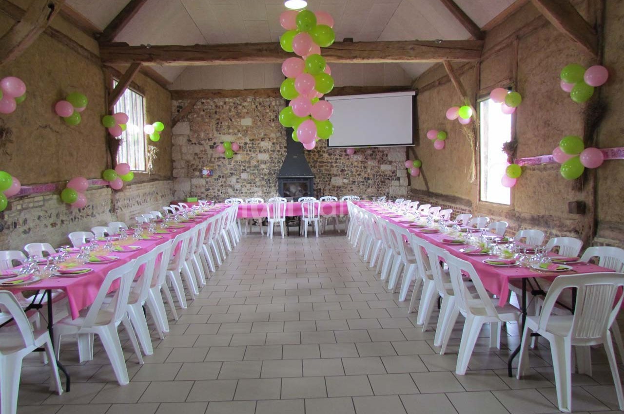 Salle de réception décorée avec des ballons roses et verts, tables alignées avec nappes roses et chaises blanches.