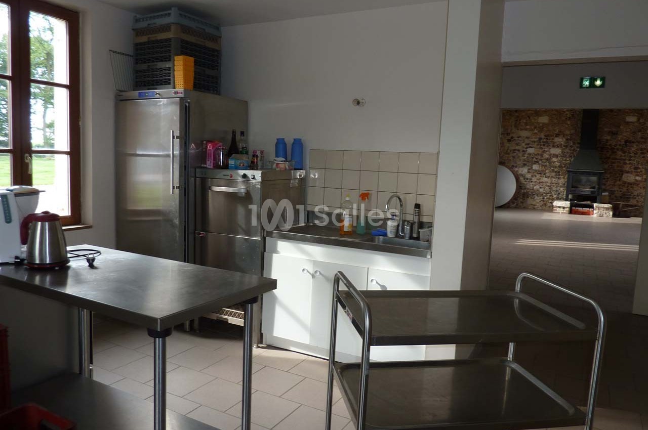 Cuisine professionnelle avec évier, plans de travail en inox, étagères et chariot, éclairée par une fenêtre.