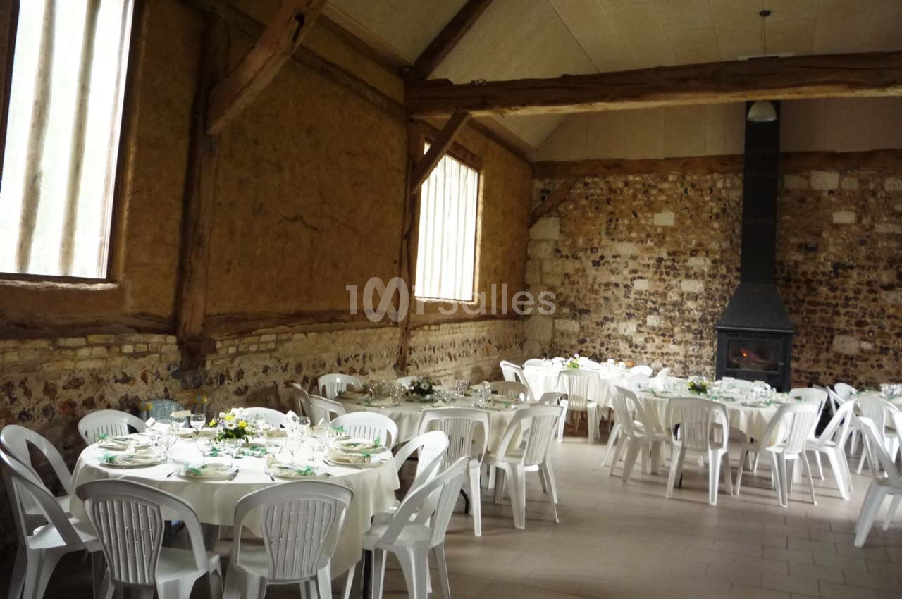 Salle rustique avec murs en pierre et poutres apparentes, tables rondes dressées avec nappes blanches et chaises en…
