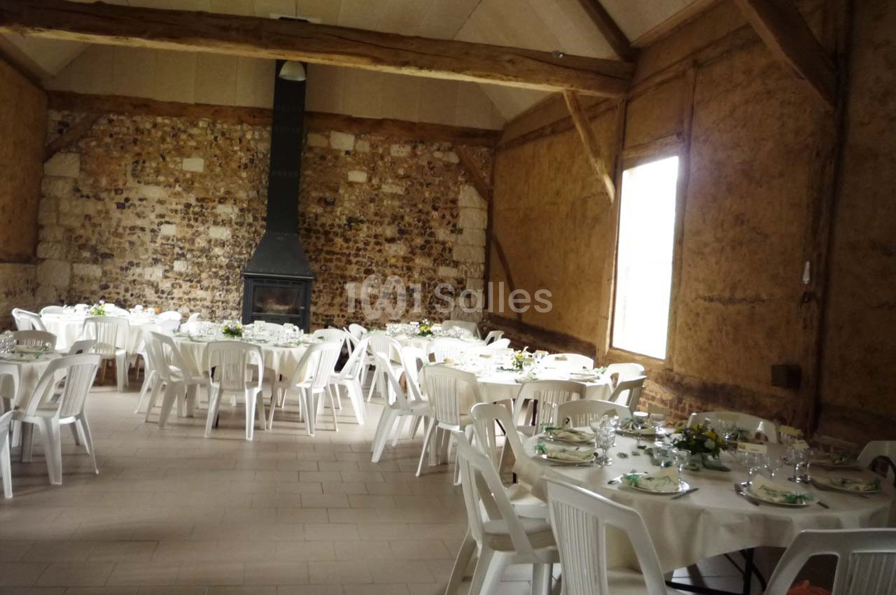 Salle rustique avec murs en pierre et poutres apparentes, tables rondes dressées avec nappes blanches et chaises en…