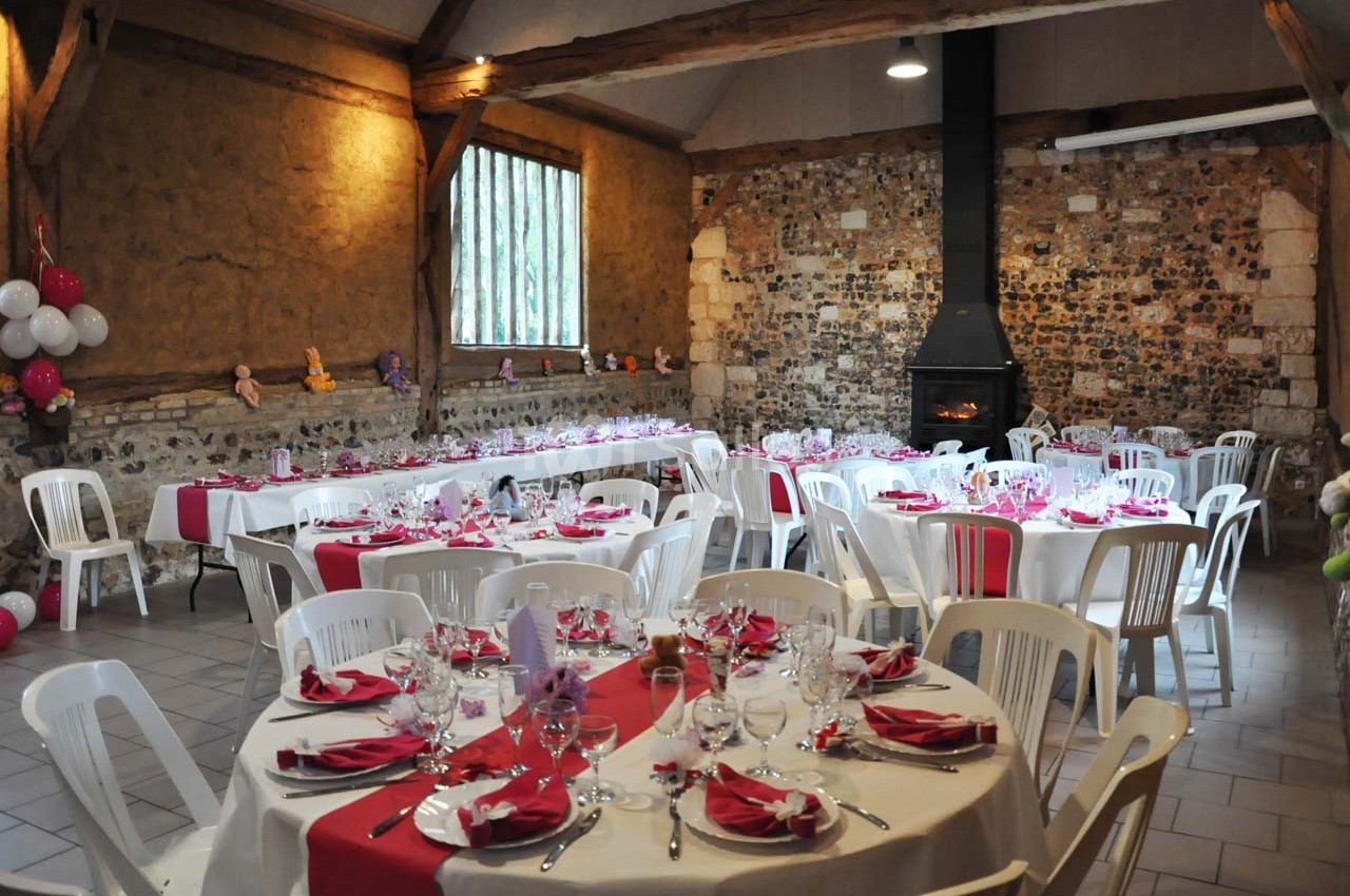 Salle de réception rustique décorée pour un événement, avec tables rondes dressées et nappes blanches et rouges.