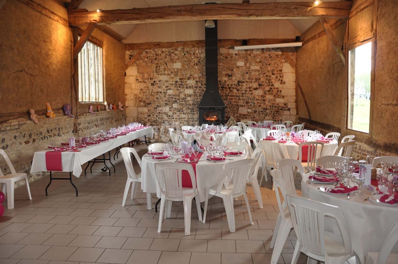 Salle rustique décorée pour un événement, avec tables dressées, nappes blanches et accents roses, cheminée au fond.