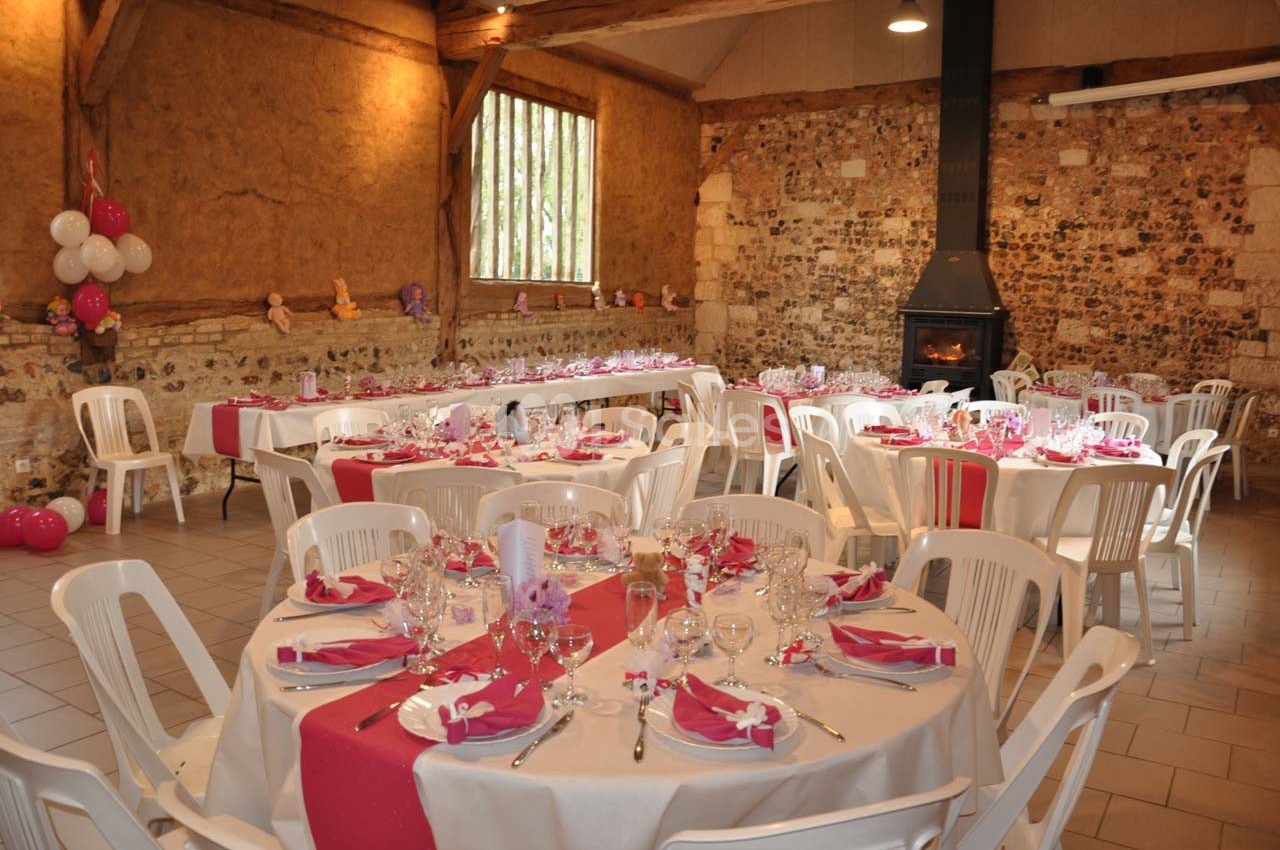 Salle décorée pour une réception, avec des tables rondes dressées, nappes blanches et chemins de table roses.
