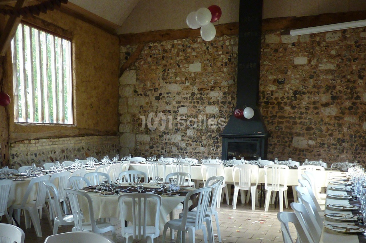 Salle rustique avec murs en pierre, tables dressées avec nappes blanches et chaises en plastique, décorée de ballons.